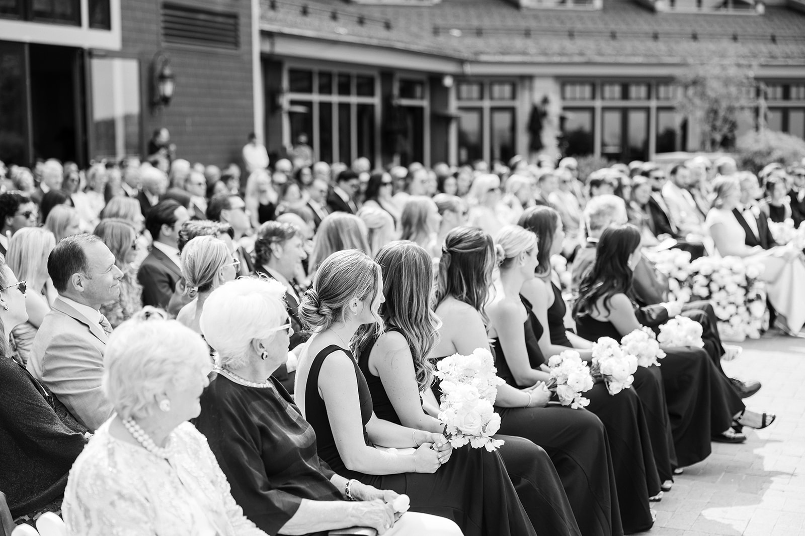 Black and white photo of guests and bridesmaids seated in the front rows during a ceremony, bouquets held in their laps