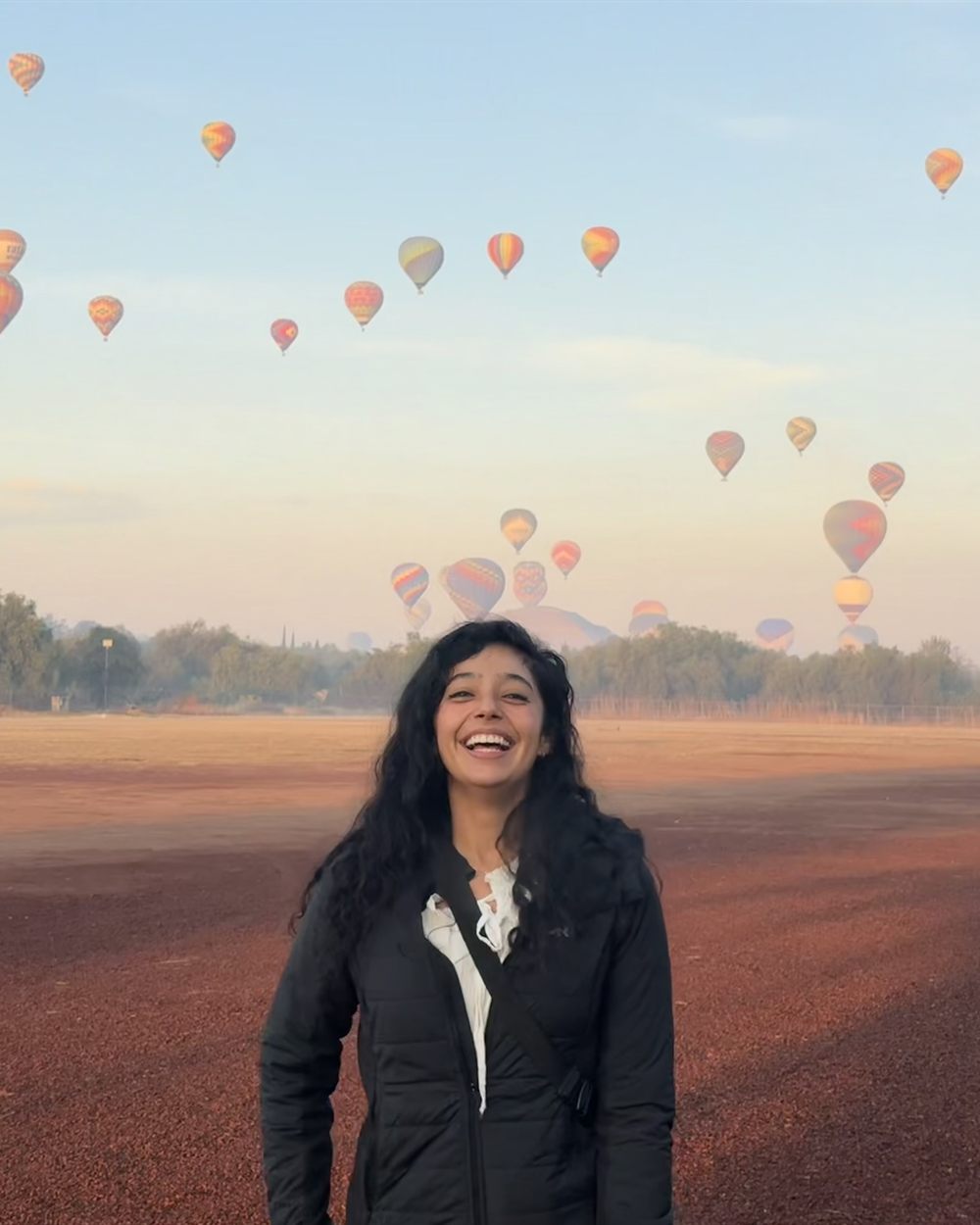 Dani Vargas laughing in an open field at dawn, with dozens of hot air balloons rising in the sky behind her