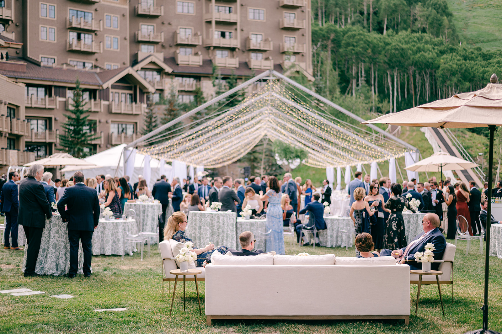 Guests at an outdoor cocktail hour under a canopy of string lights, with lounge furniture on the lawn and a mountain lodge behind them