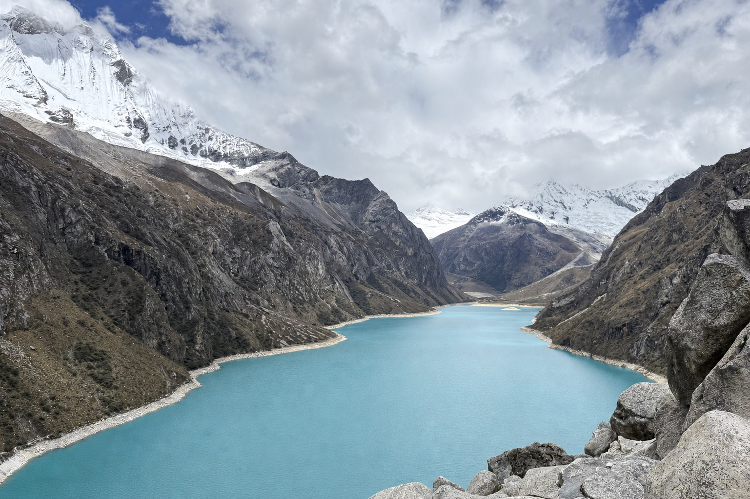 A turquoise mountain lake beneath snow-capped peaks and a cloud-filled sky