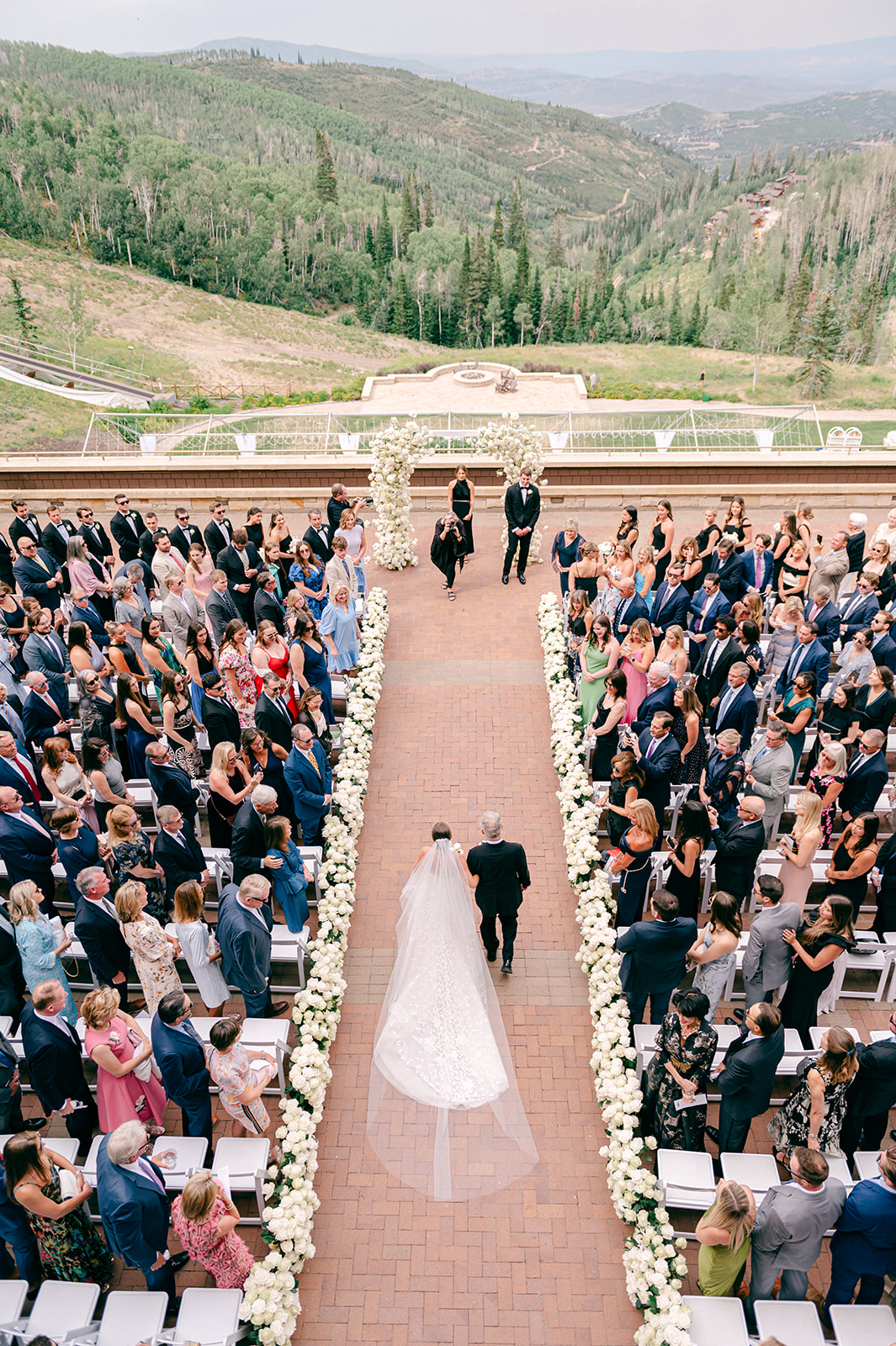 An overhead view of an outdoor mountain ceremony, the bride walking a rose-lined aisle with her father as guests stand on both sides