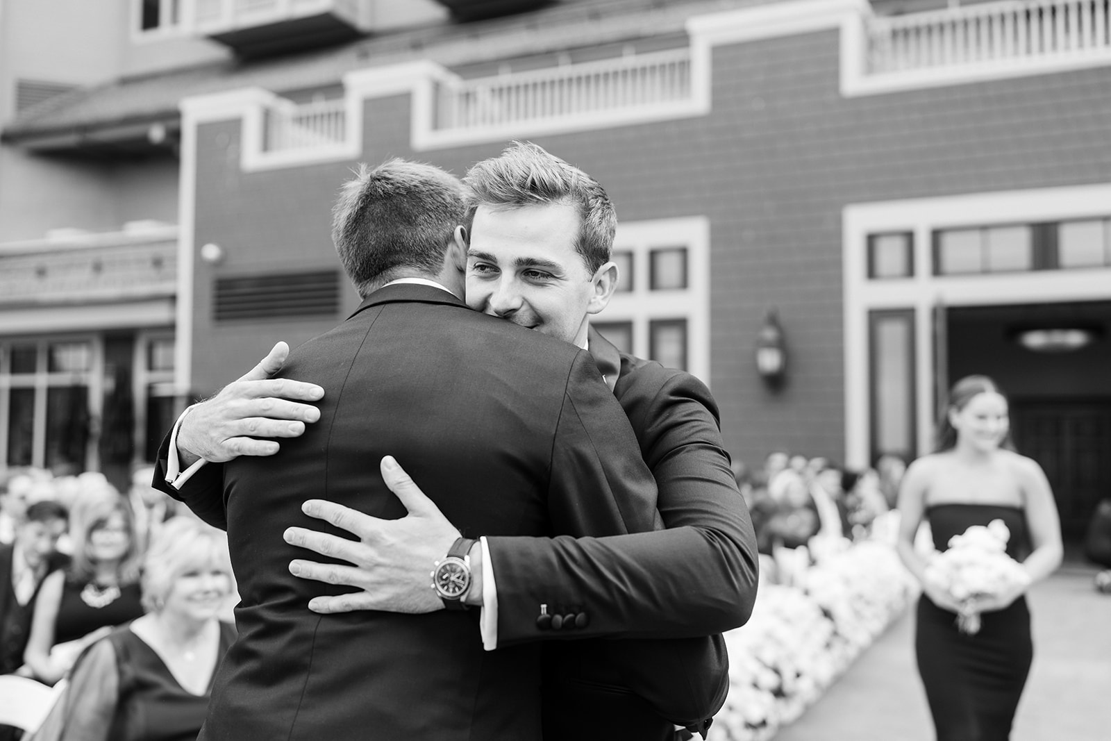 Black and white photo of a groom embracing a guest at the end of the aisle, smiling over his shoulder as the bridesmaids come down behind them