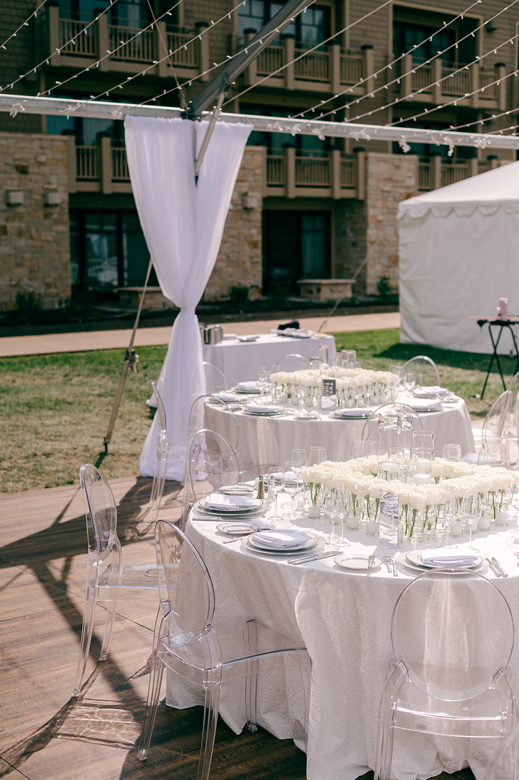 Round reception tables set with white linens, clear ghost chairs, and low white rose arrangements beneath a canopy of string lights